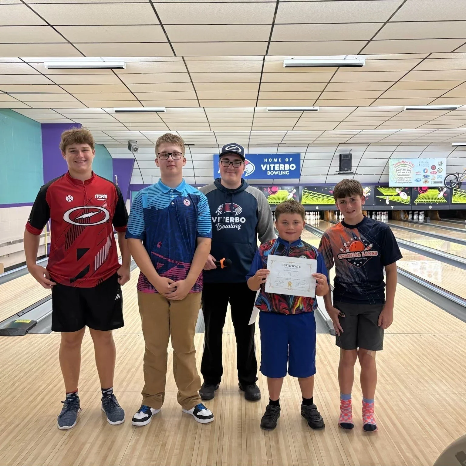 A group of five boys standing at a bowling alley, with one boy holding a certificate. The background shows bowling lanes and a sign that reads 'Home of Viterbo Bowling'.