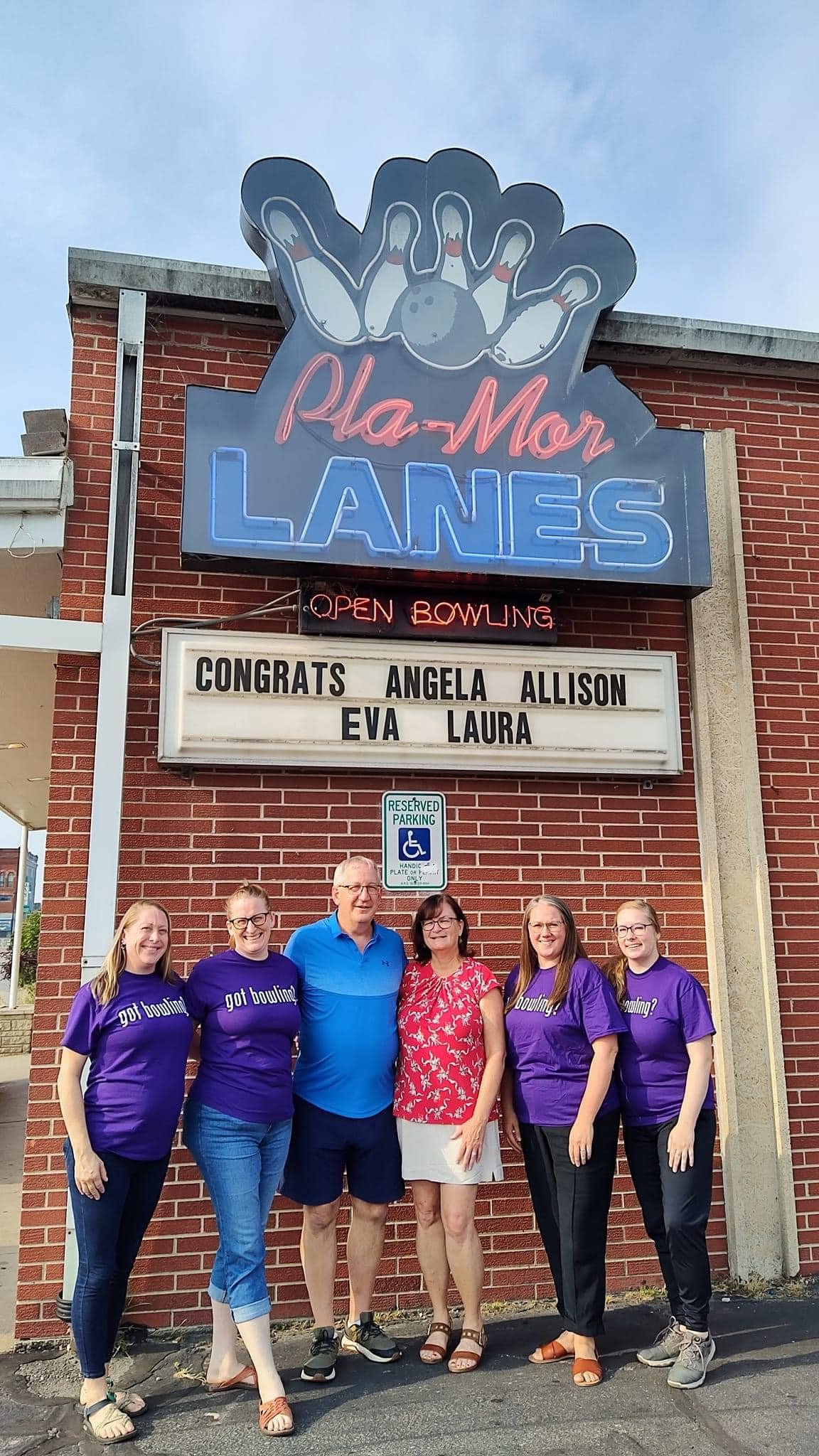A group of six people standing in front of a brick bowling alley sign that reads "Pla-Mor Lanes." Overhead neon sign says "Open Bowling." The group includes four women wearing purple shirts with white text, a man in a blue shirt, and a woman in a red top with white shorts. They are all smiling, standing on the pavement outside the bowling alley, under a parked reserved parking sign.