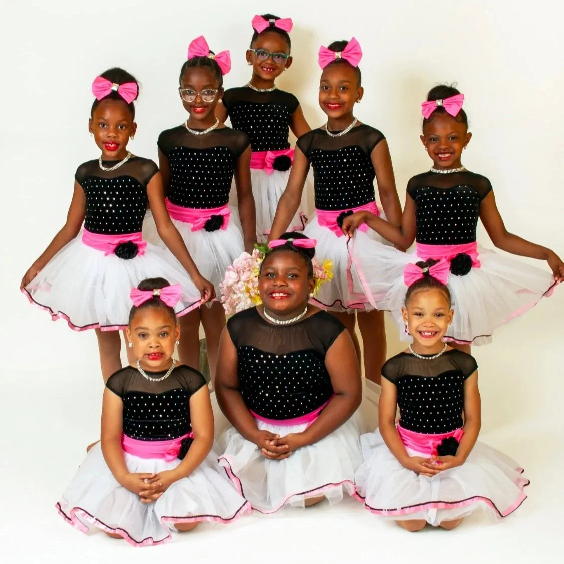 Group of nine young girls dressed in matching black, pink, and white dance costumes, smiling for a photo against a plain white background.