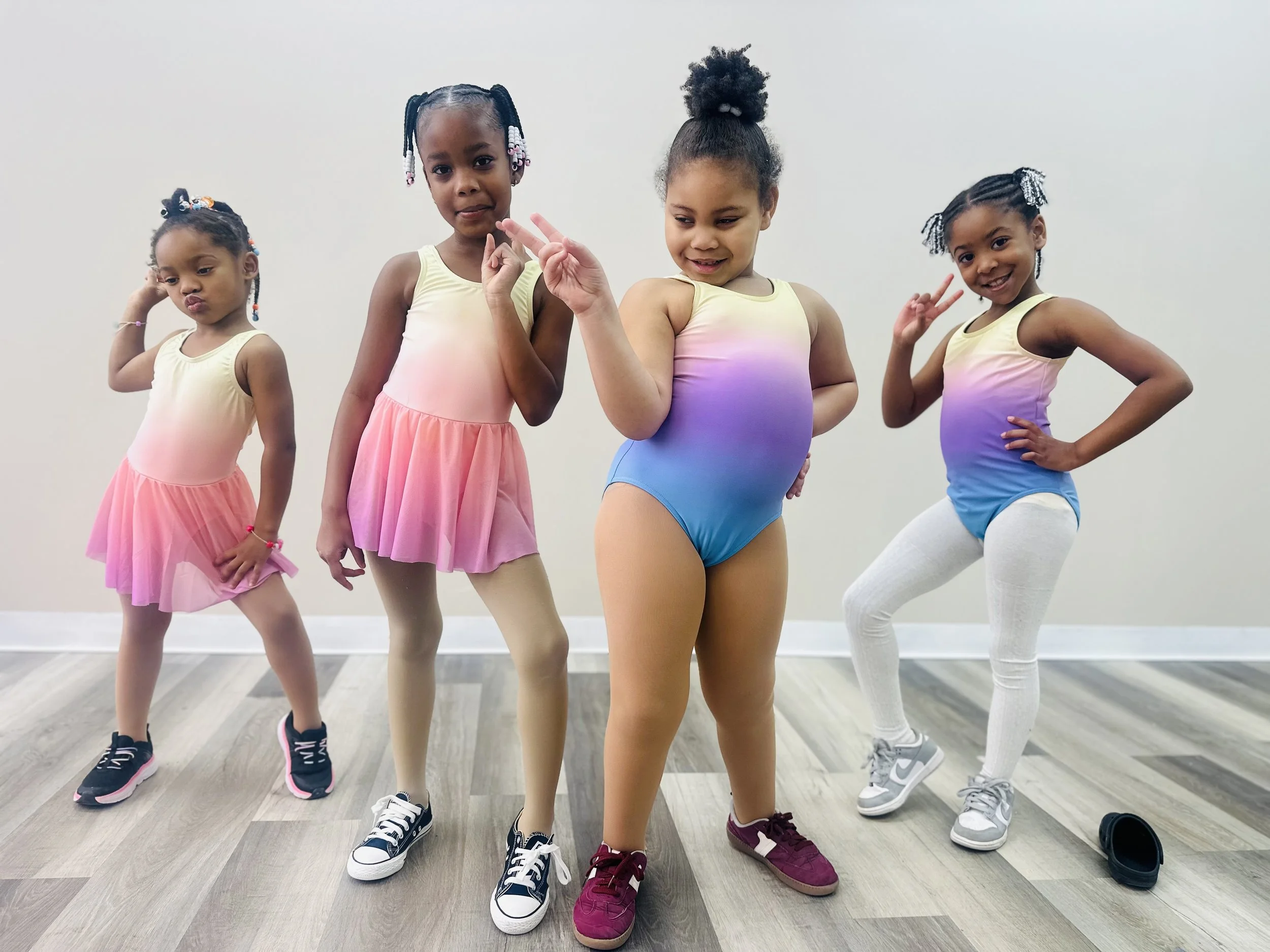 Four young girls in colorful gradient dance leotards and sneakers striking playful poses in a dance studio.