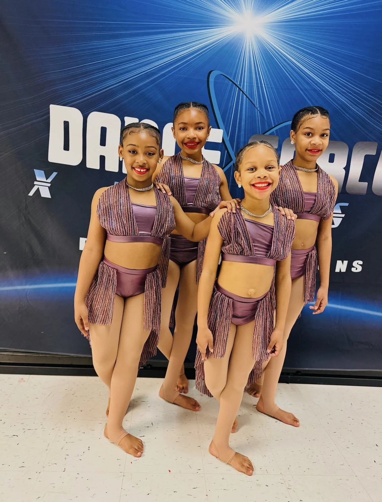 Five young girls in purple and pink dance costumes posing together at a dance competition venue.