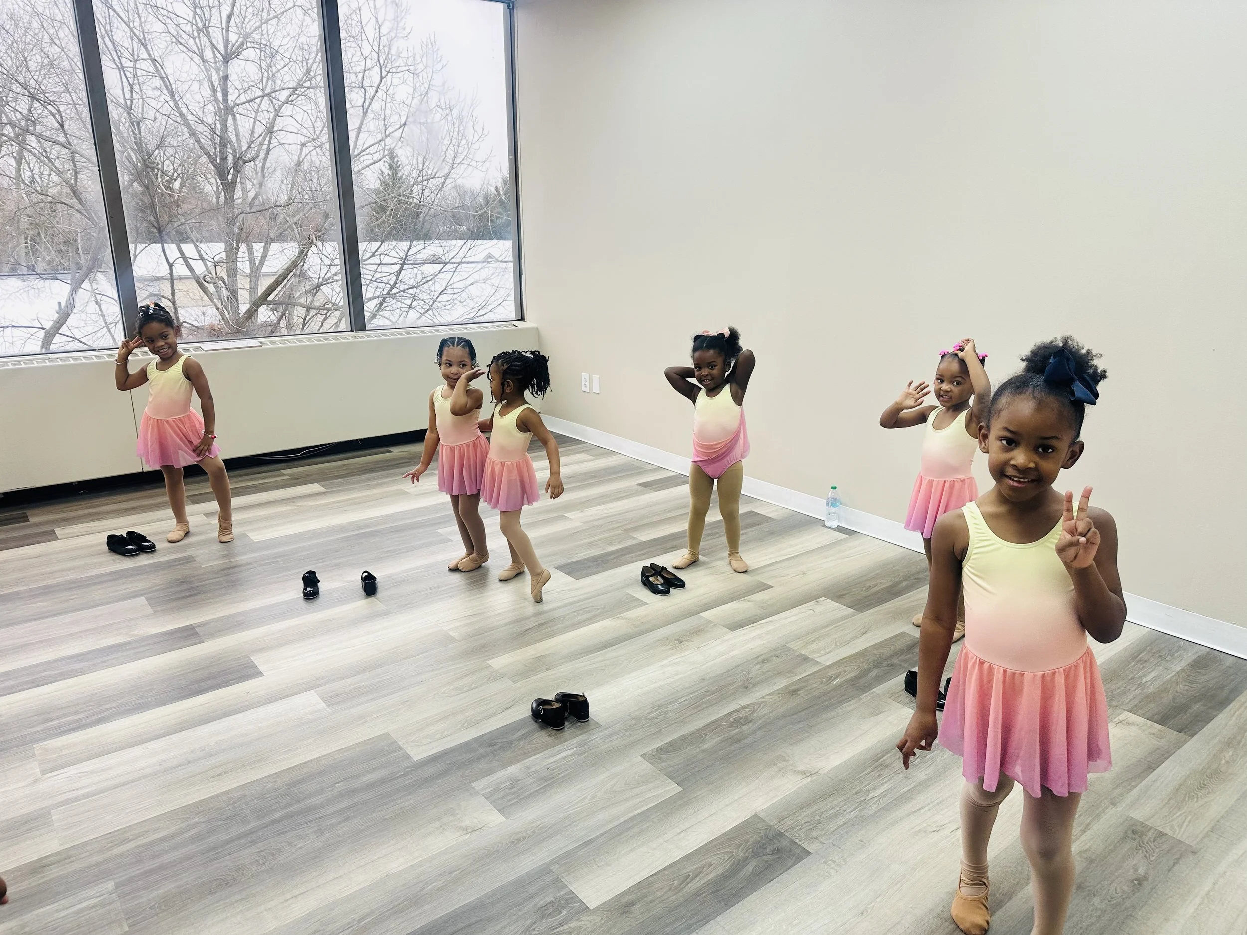 Six young girls dressed in pink ballet dresses and tights standing in a dance studio with a large window showing a snowy landscape outside. Some girls are posing while others are adjusting their hair. Shoes are on the floor.