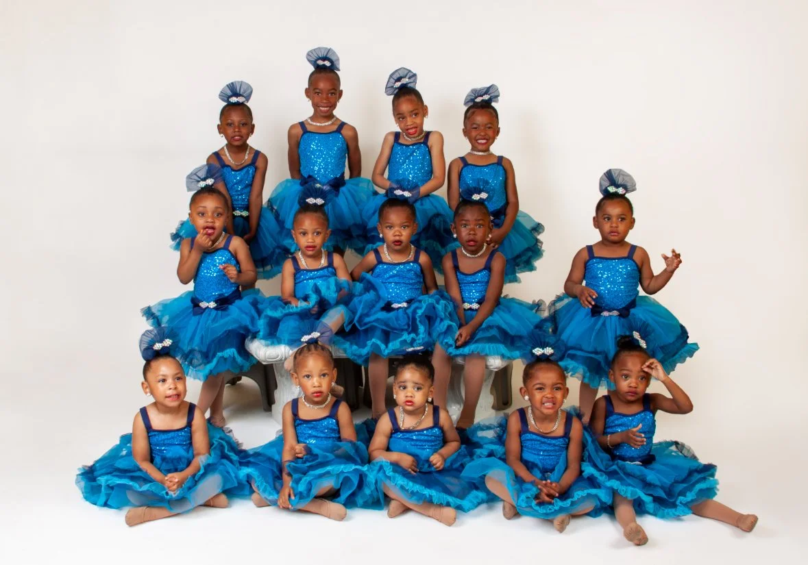 Group of young girls in matching blue dance costumes with frilly skirts and bows, posing together in a studio.