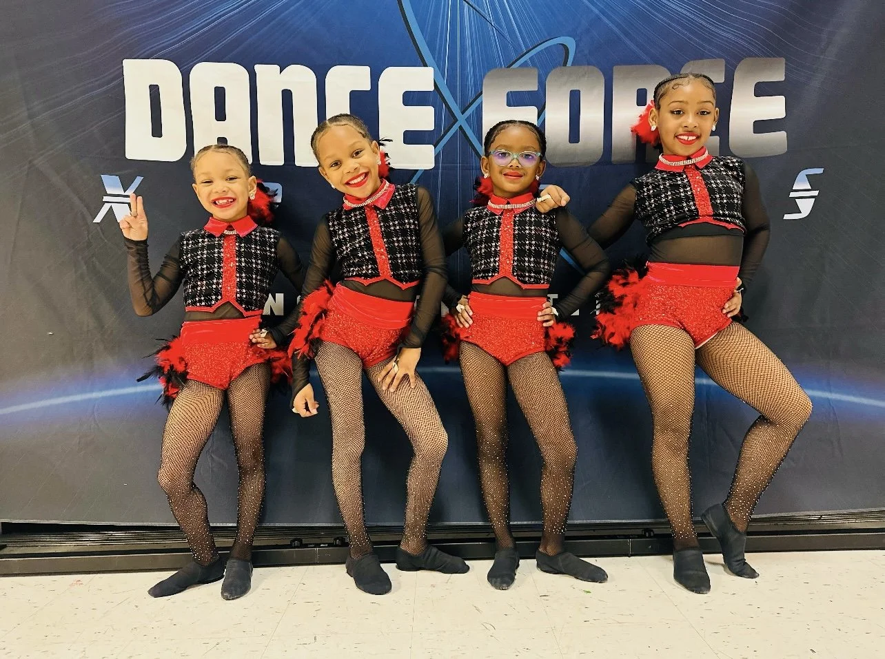 Four young girls in dance costumes posing in front of a backdrop with the words 'Dance Force'. They are wearing black, red, and sparkly costumes with fishnet stockings, and their hair is styled with red accessories.