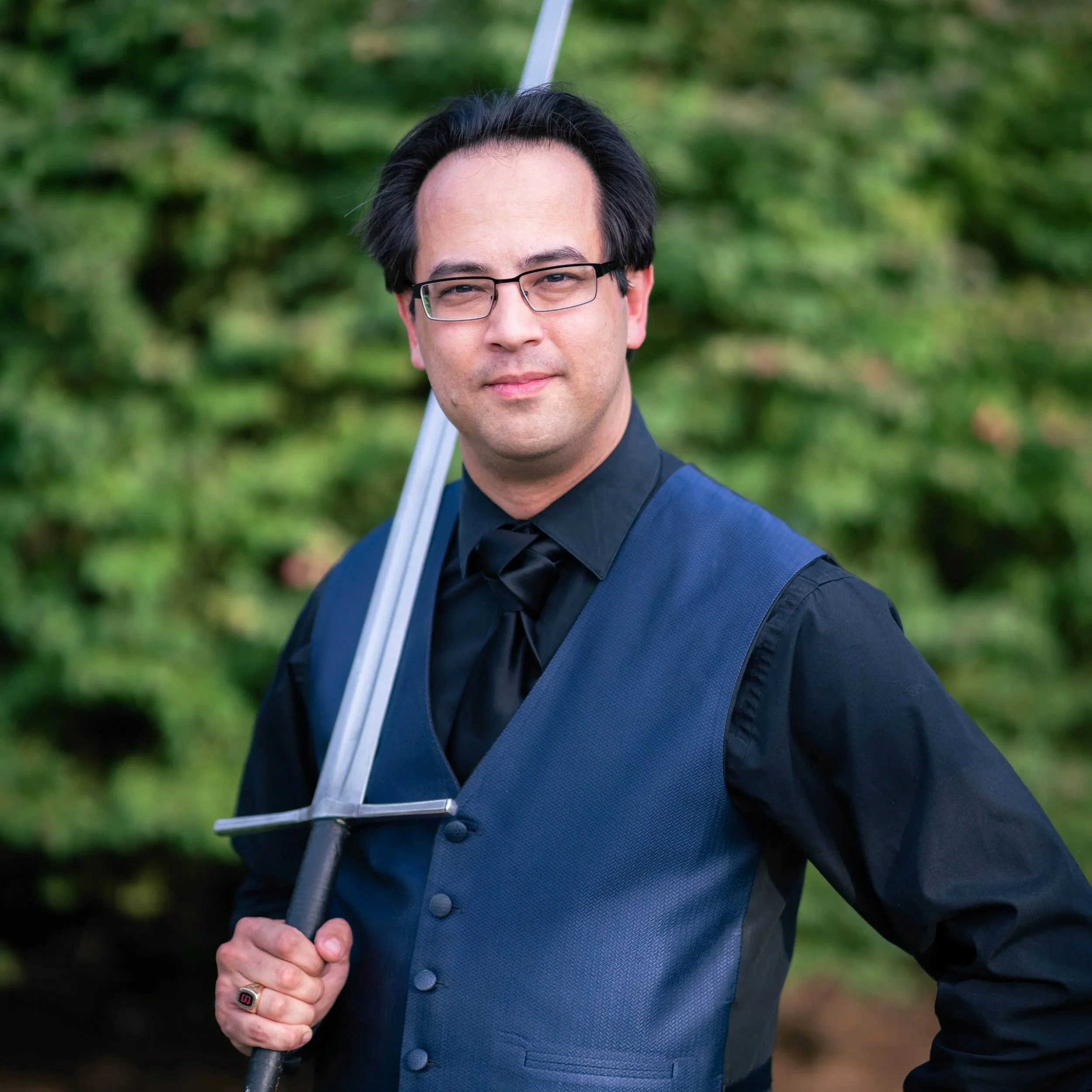 A man dressed in a dark blue vest and black shirt holding a sword outdoors, with trees and greenery in the background.