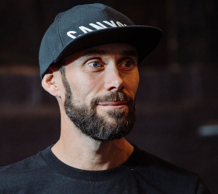 A man wearing a black baseball cap and black T-shirt, with a beard, in an indoor setting.