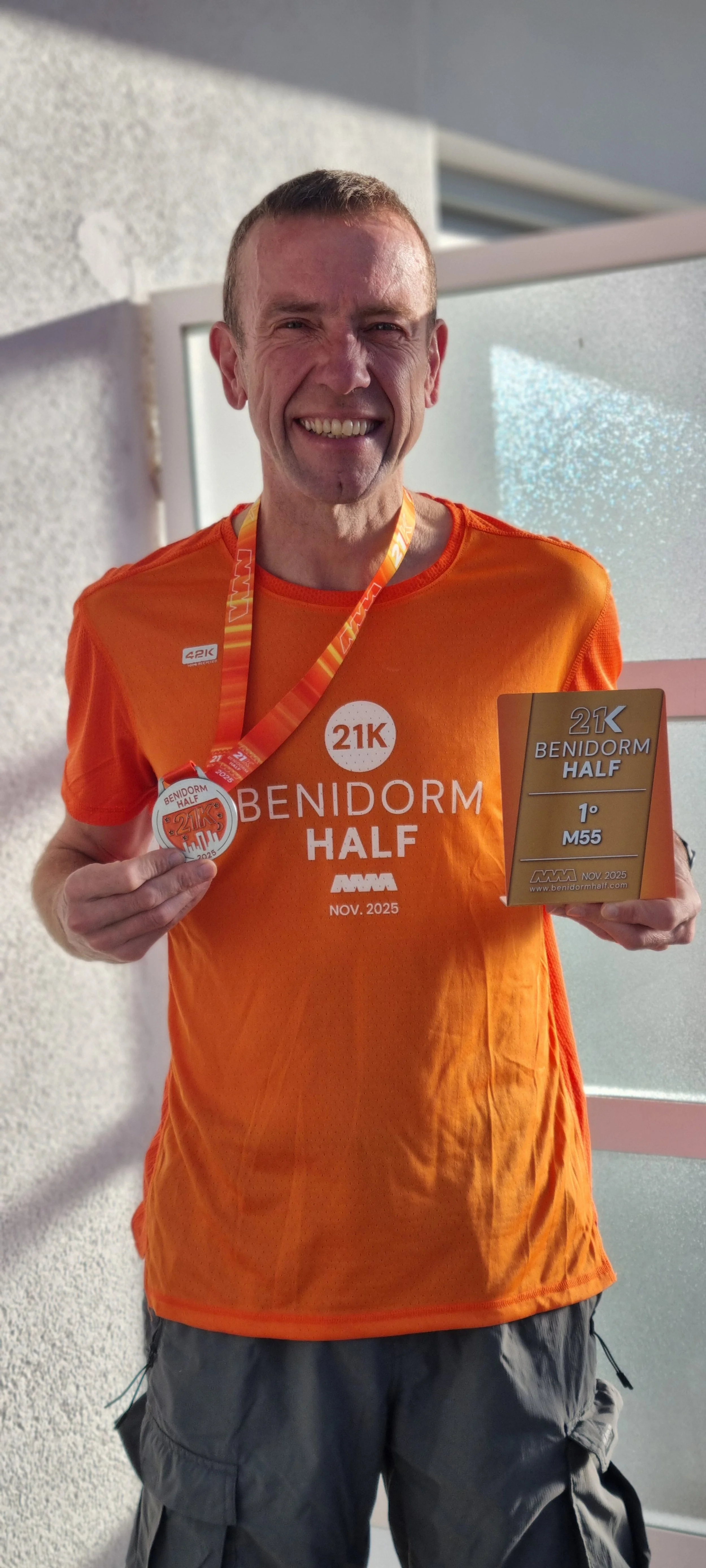 A man smiling and holding a medal and a plaque after completing the Benidorm Half marathon in November 2025, wearing an orange shirt with the event's logo.