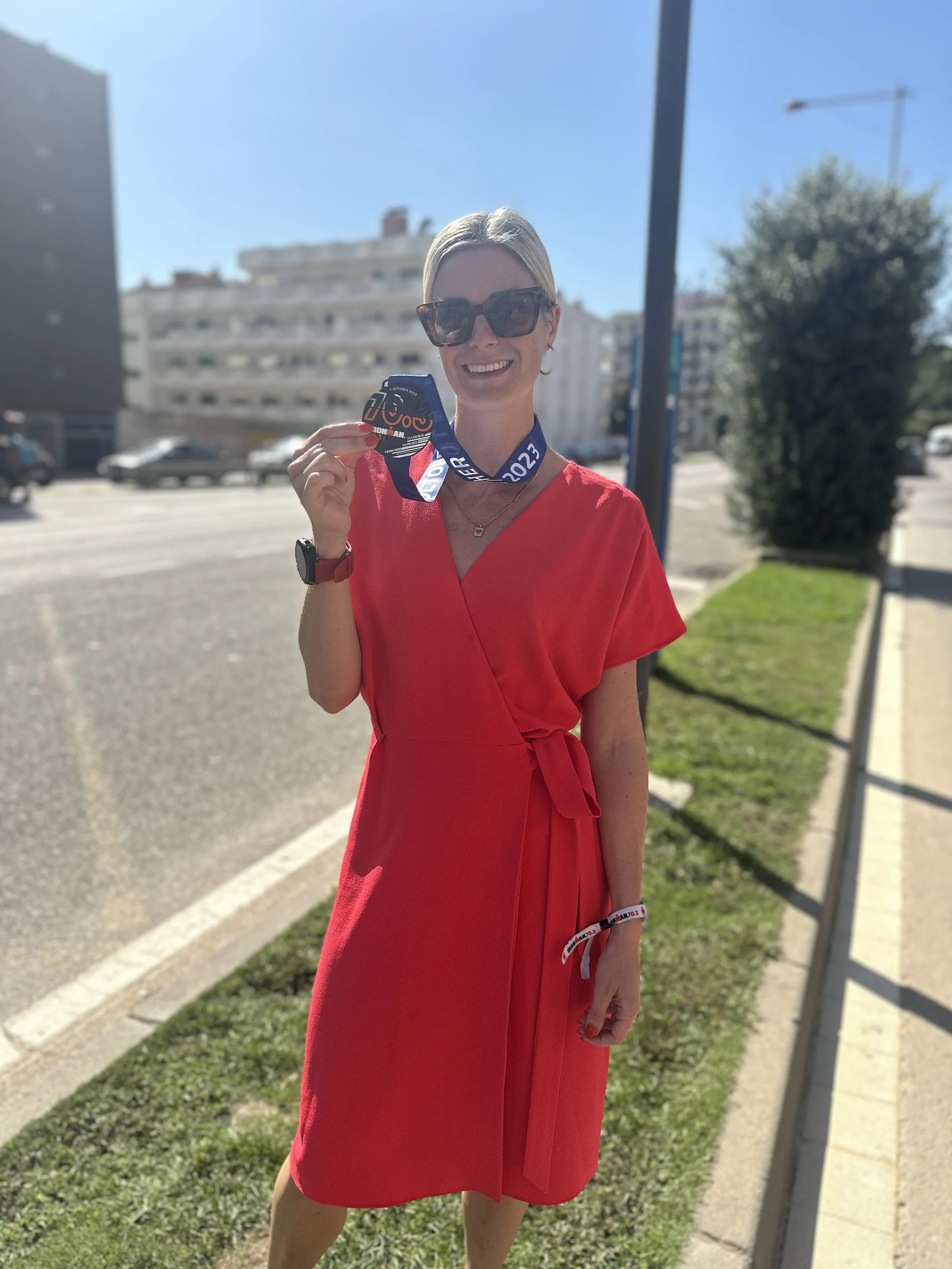 A woman smiling and holding a finisher medal from a 2023 race, standing on a sidewalk with buildings and cars in the background.