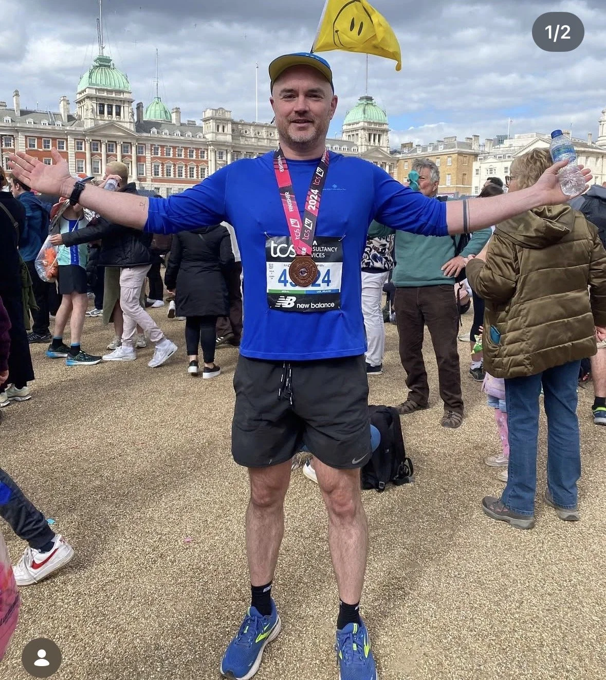 Man wearing blue long sleeve shirt, black shorts, and running shoes standing with arms open during a marathon event, with a finishers medal around his neck, a yellow flag with smiley face, and race bib number 4624. Crowds of people and historic buildings are in the background.