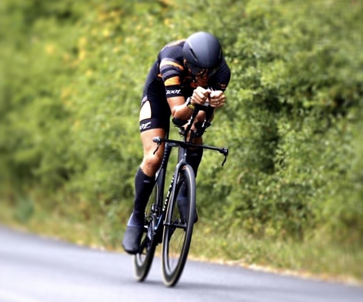 A cyclist in black and orange sports gear and helmet, riding a black racing bicycle on a rural road with green foliage in the background.