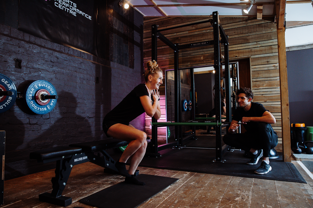 A woman seated on a workout bench, laughing and clasping her hands in front of her chest, while a trainer crouches nearby, smiling and holding a drink, in a gym with wooden and brick walls and gym equipment.