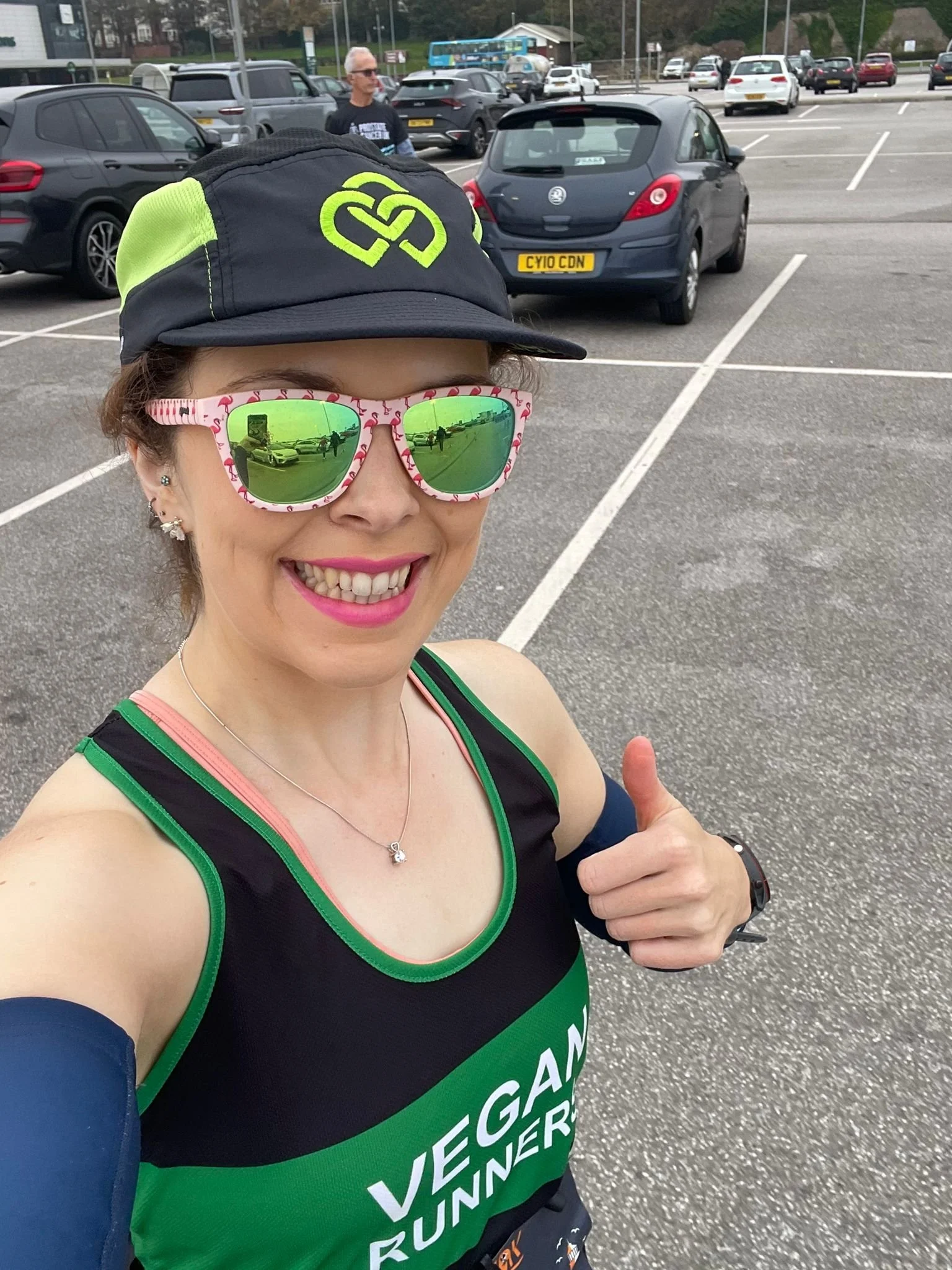 A woman taking a selfie in a parking lot, dressed in athletic gear with « VEGAN RUNNER » printed on her shirt, wearing sunglasses, a cap, and giving a thumbs-up.