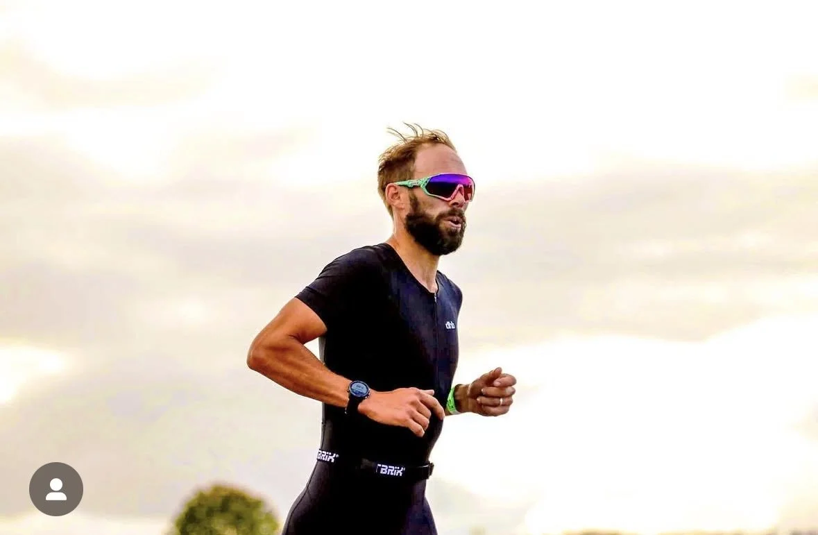 Man running outdoors wearing sunglasses, black athletic shirt, and black shorts, with a cloudy sky in the background.