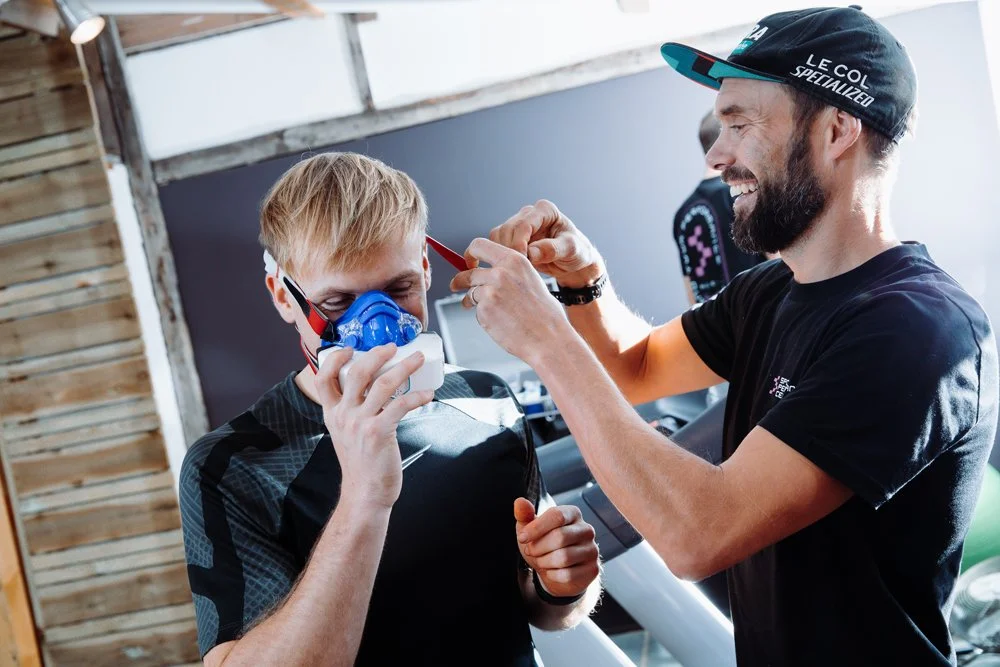 A man helping a young boy put on a mask for a biking activity, both smiling and indoors at a gym or fitness center.