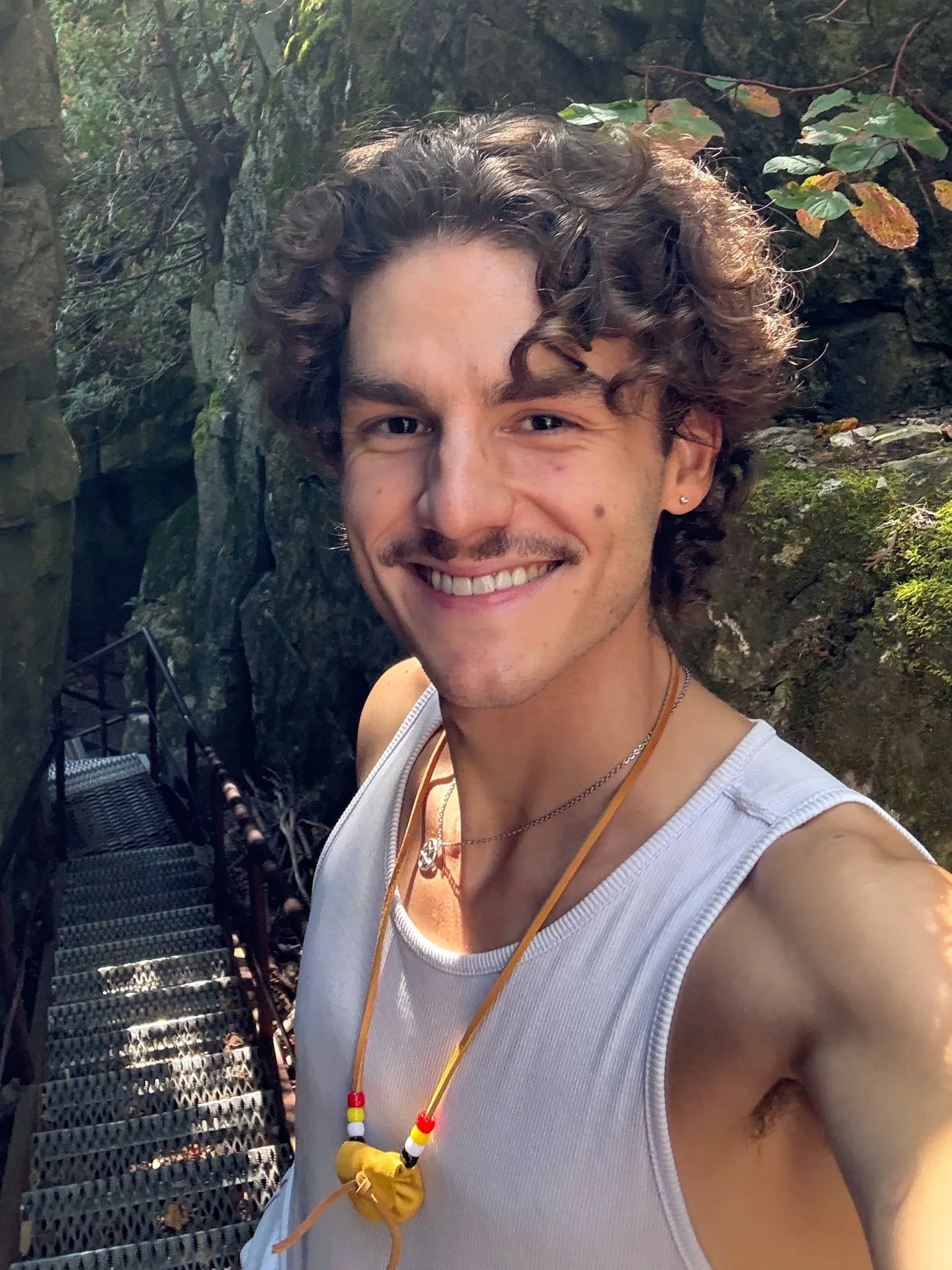 A young man with curly hair and a mustache smiling, wearing a white tank top and a necklace with beads, taking a selfie outdoors in front of rocks and greenery.
