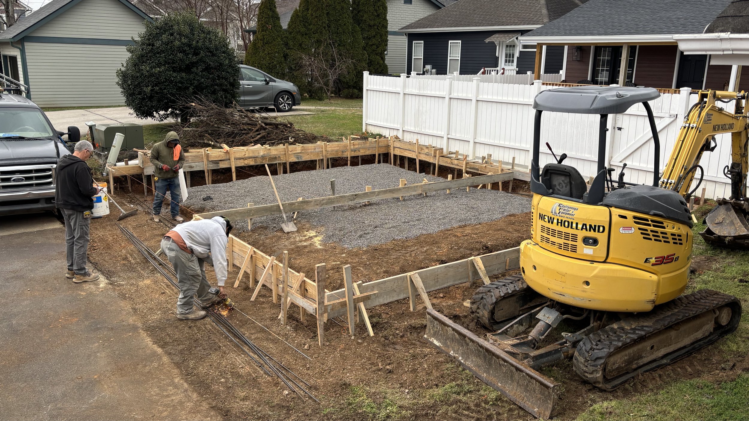 Construction workers are building a foundation for a new structure in a residential backyard. There is a yellow mini excavator labeled 'New Holland' on the right, and wooden framing around the construction area. Some workers are welding, and others are working with tools. A white fence separates the yard from neighboring houses, and several cars are parked nearby.