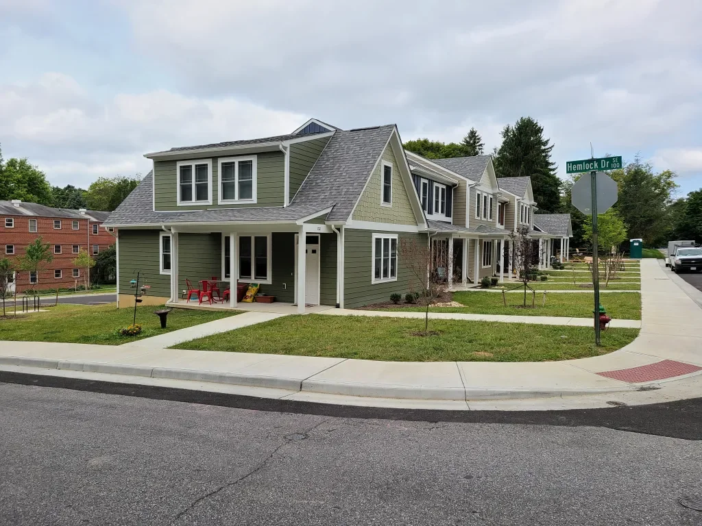 A row of newly built green and beige townhouses with porches, situated on a corner lot at Hemlock Drive Southeast. The sidewalks and lawns are well-maintained with young trees and small garden beds.