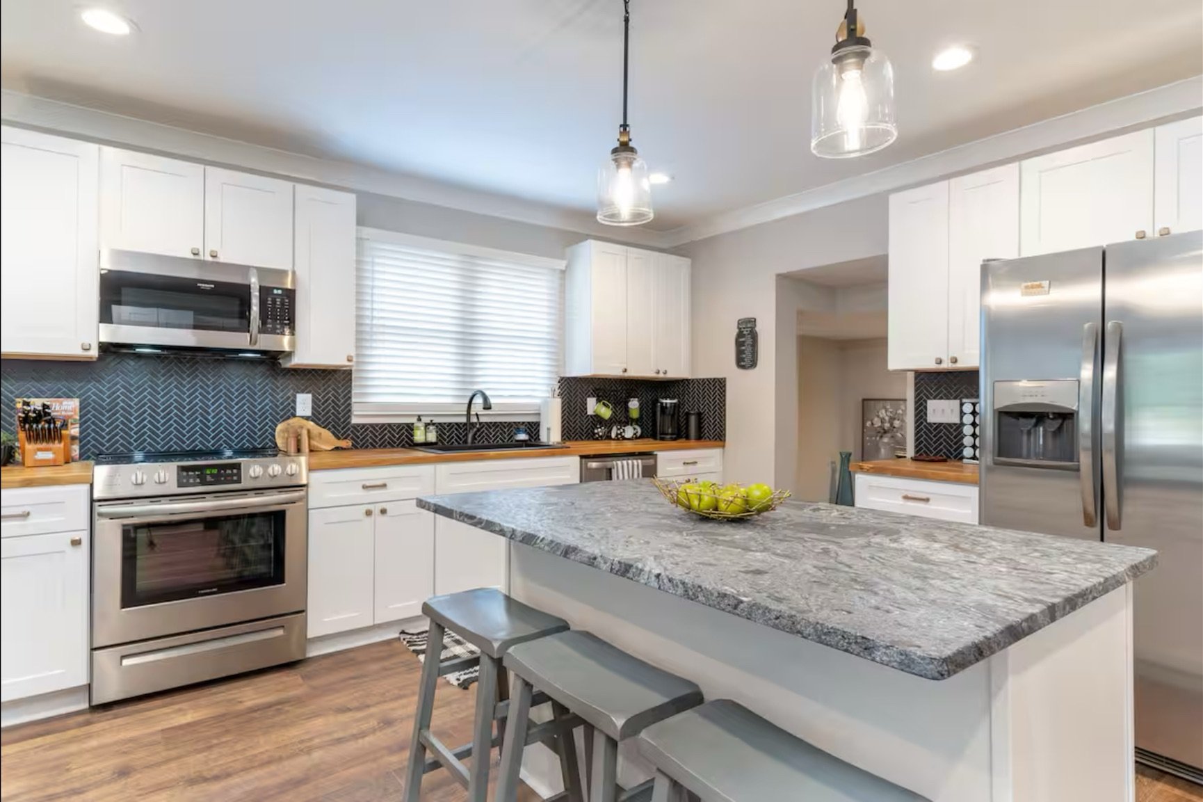 Modern kitchen with white cabinets, stainless steel appliances, and a granite island with three bar stools. Black backsplash, wooden countertops, and a bowl of green apples on the island.