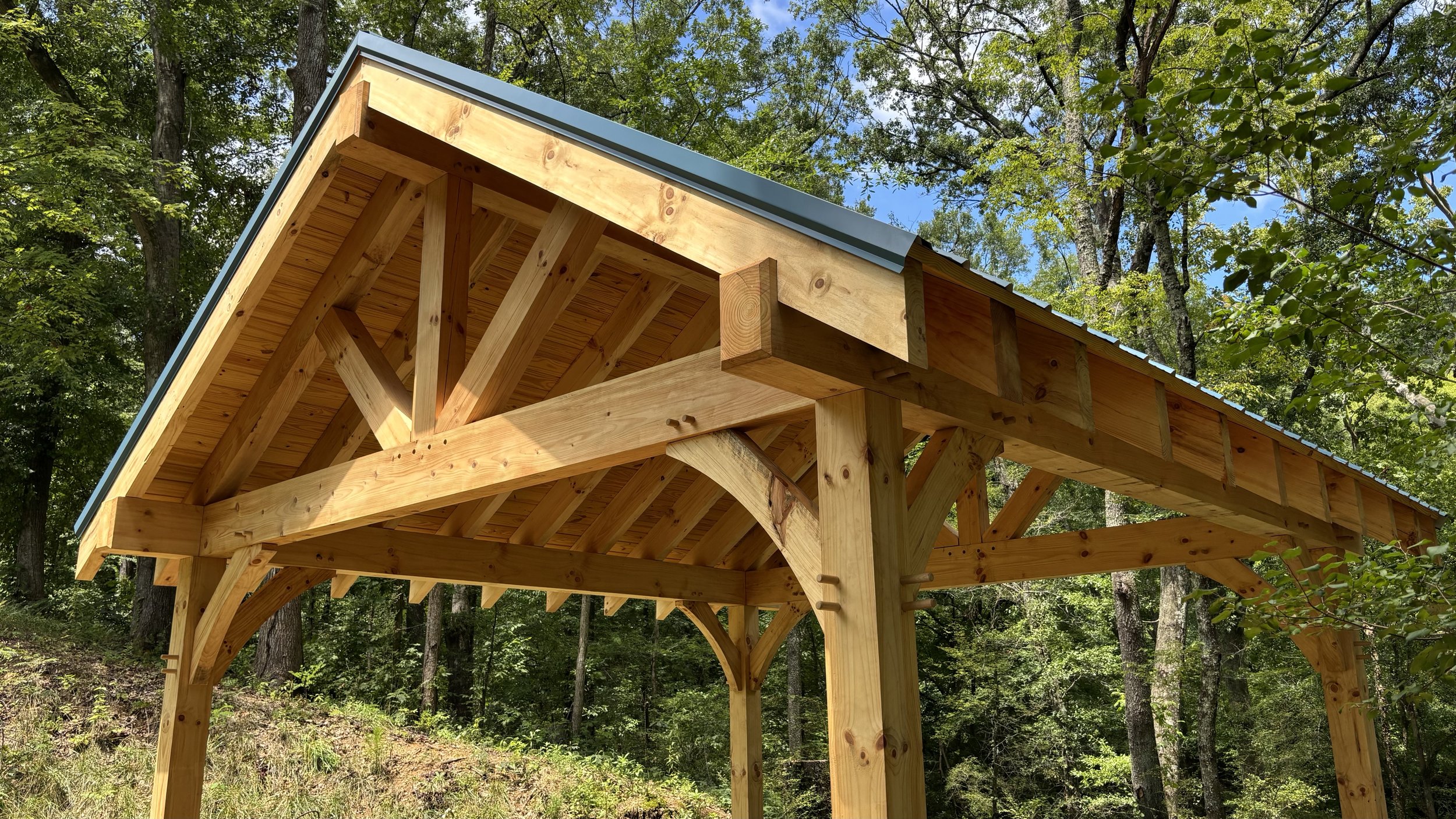 Close-up of a wooden deck under construction in a forested area, showing the wooden framework and beams with trees and sky in the background.