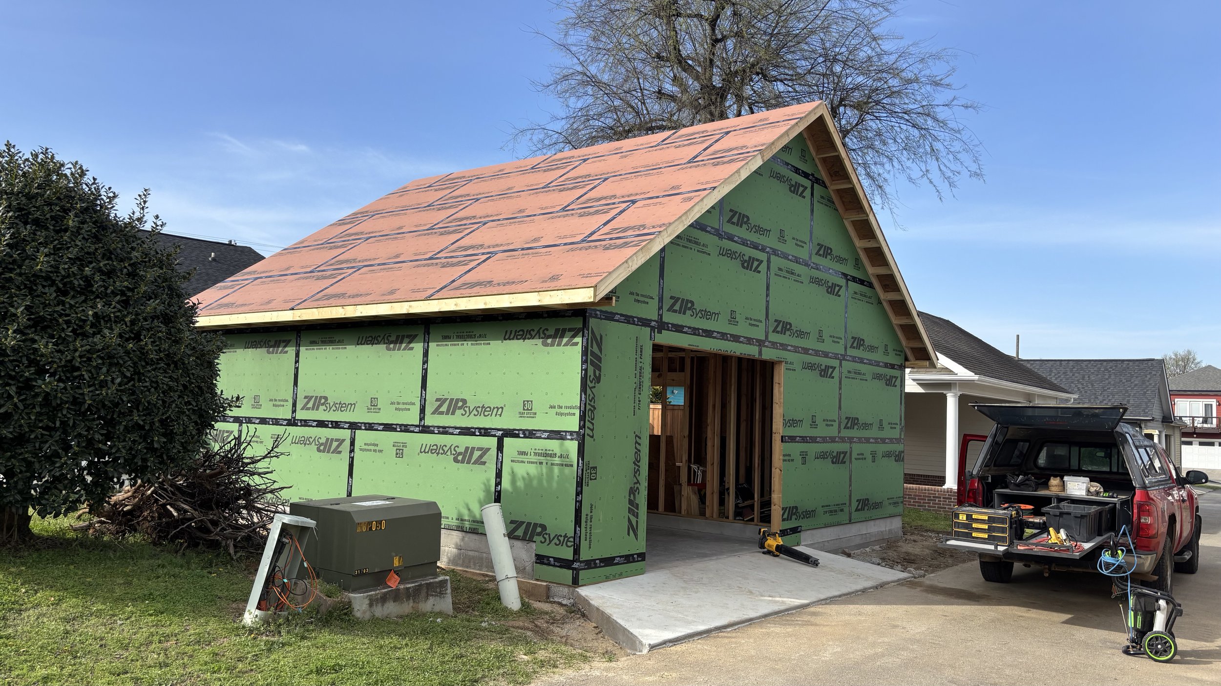 A house under construction with green sheathing and a pitched roof, partially built, next to other completed houses in a neighborhood with a blue sky.