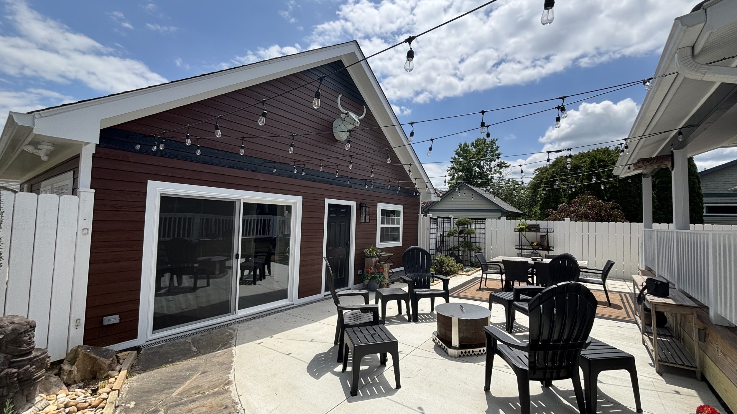 Backyard patio area with black chairs and tables, potted plants, string lights overhead, and a red house with a black door and sliding glass door. Fencing surrounds the area under a partly cloudy sky.