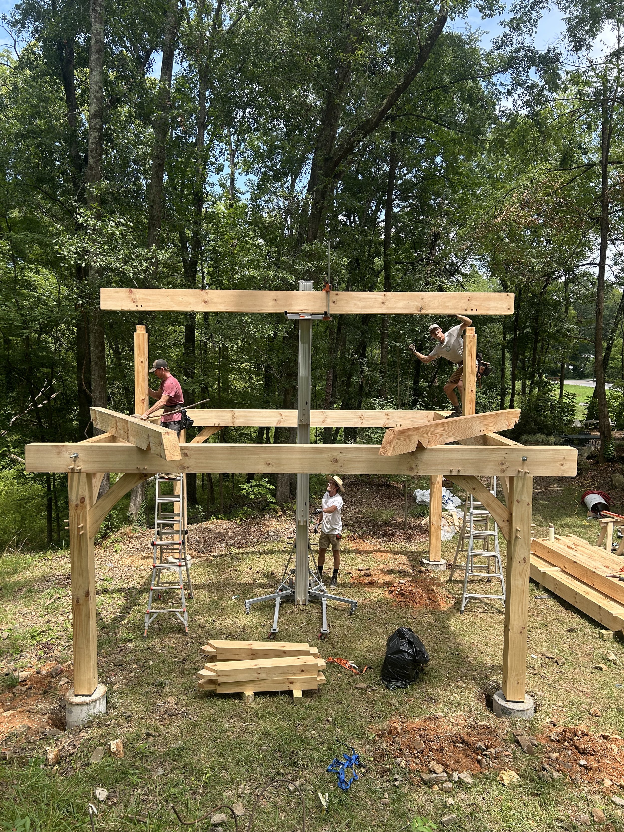 Three men building a wooden structure outdoors in a wooded area. The structure has vertical and horizontal beams, with two ladders and construction tools nearby.