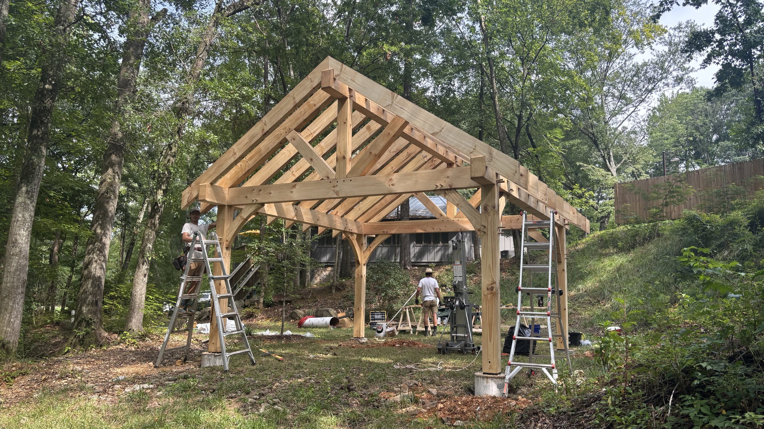 Construction of a wooden pavilion in a wooded area, with workers and tools present.