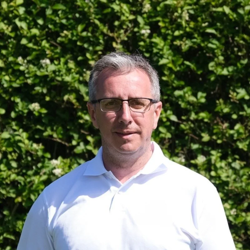 A man with glasses and gray hair standing outdoors in front of a green leafy background, wearing a white collared shirt.