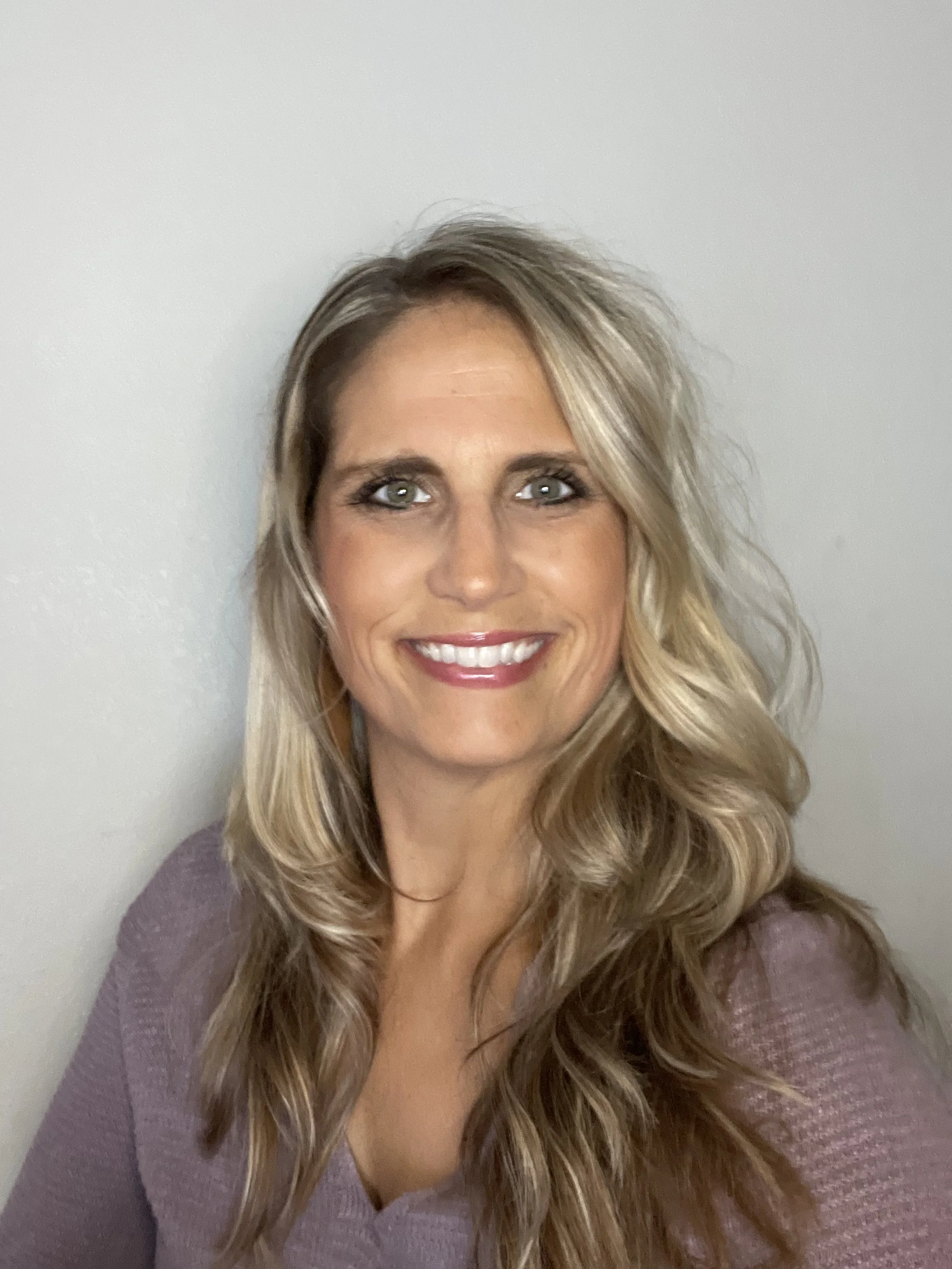 A woman with long, wavy blonde hair and green eyes smiling at the camera, wearing a purple top with a v-neck, standing against a plain light gray wall.