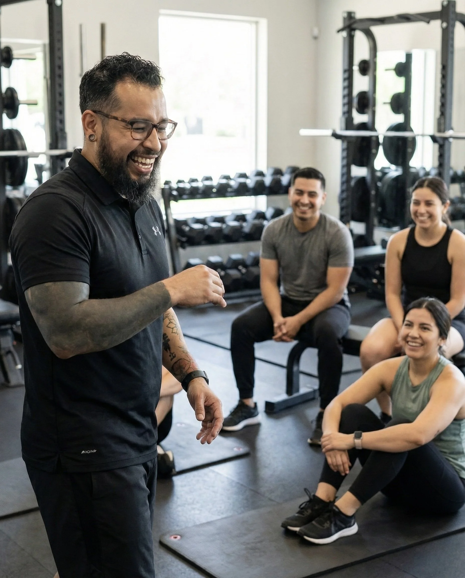 A male trainer with glasses and tattoos on his arms leading a fitness class in a gym, with three smiling women sitting on mats and benches, surrounded by gym equipment.