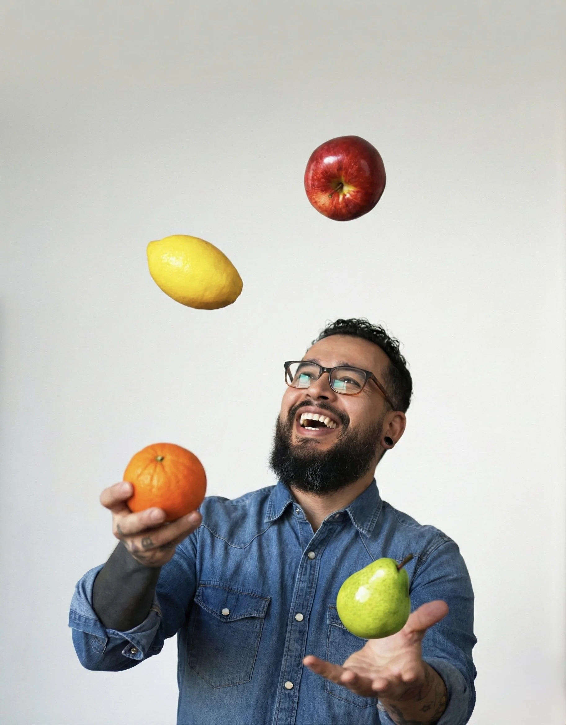 A man with a beard, glasses, and tattoos juggling a green pear, an orange, a lemon, and a red apple against a plain white background.