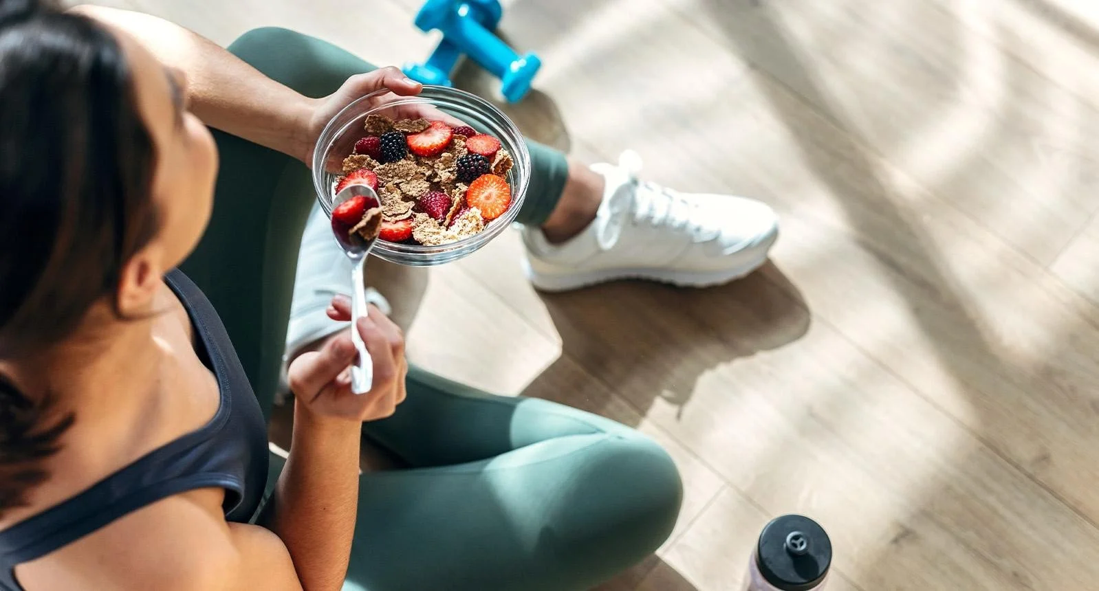 A woman sitting on a green exercise ball, holding a bowl of cereal with strawberries and blackberries, and eating with a spoon.