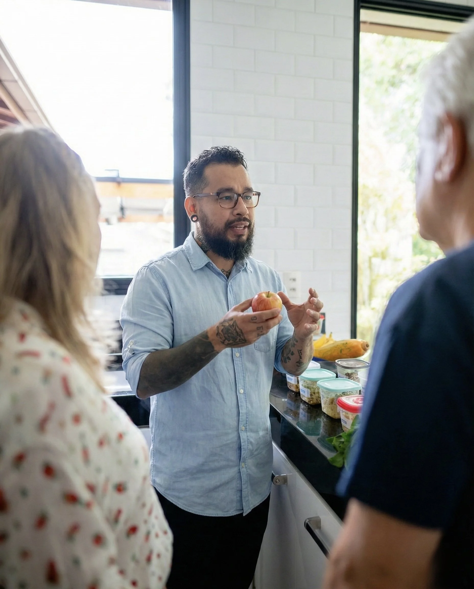 Man with glasses and tattoos explaining something about an apple to two women in a kitchen with containers of food on the counter.