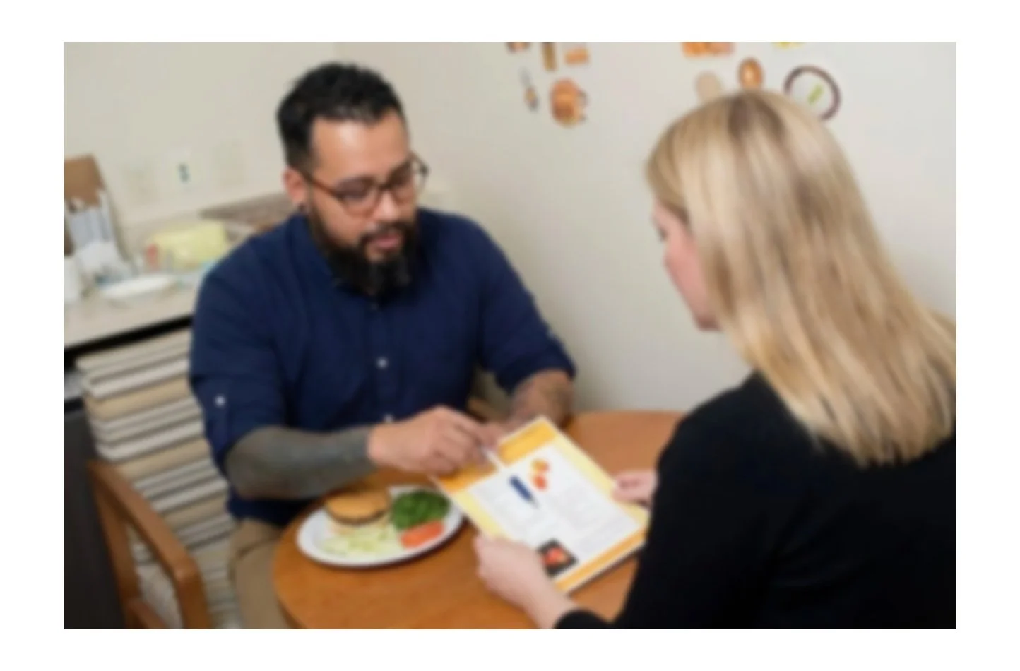 A man with glasses and a beard sitting at a table with a woman, looking at a brochure or menu. The man has a burger on a plate in front of him. They are in a casual setting, possibly a restaurant or cafe.