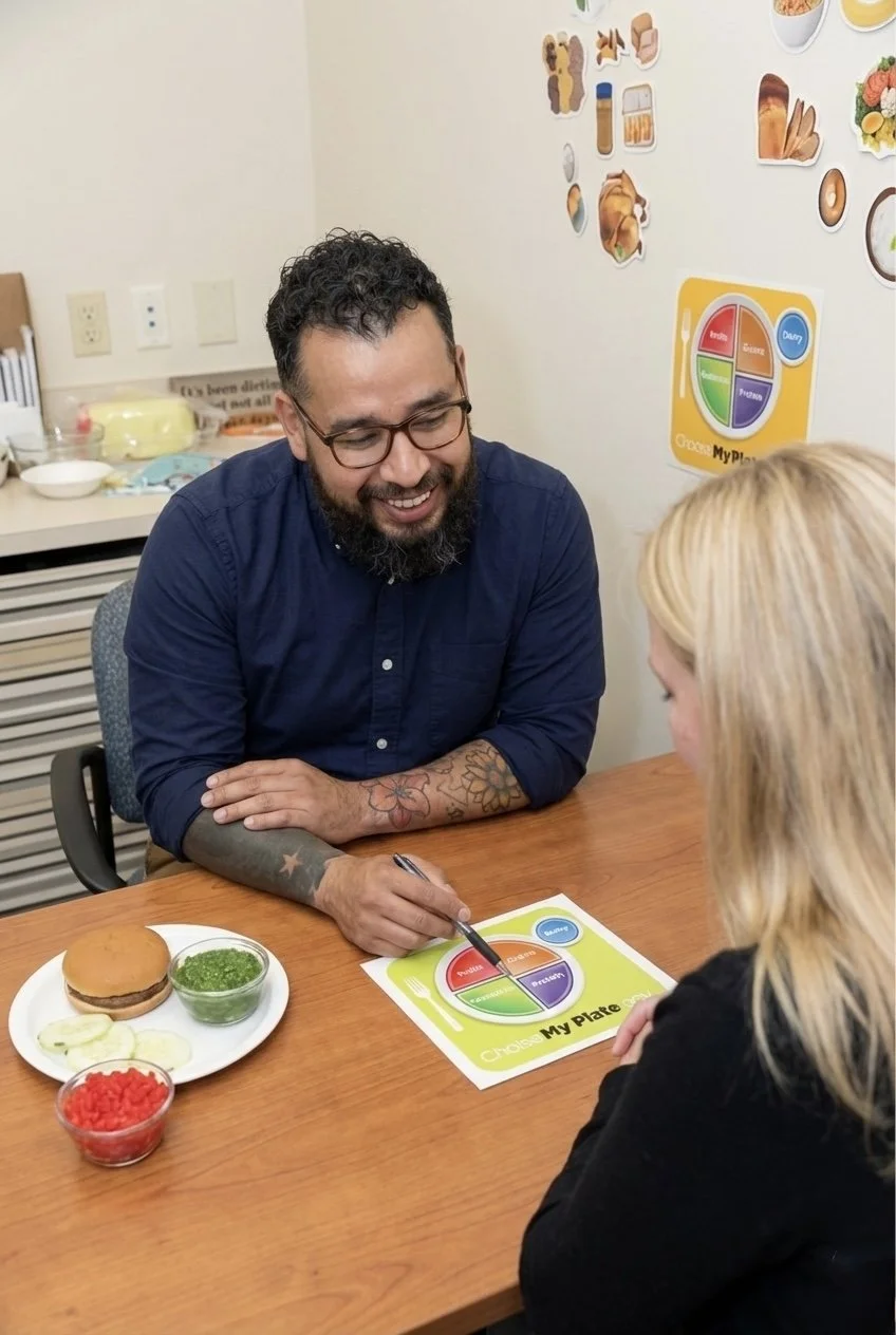 A man with glasses and a beard, smiling, sharing a meal with a woman in a counseling or therapy session. They are seated at a wooden table with a burger, a bowl of green pesto, and a small bowl of red candies. The man is pointing at a colorful plate with segments labeled 'Protein,' 'Vegetables,' 'Grains,' 'Fruits,' and 'Dairy'.