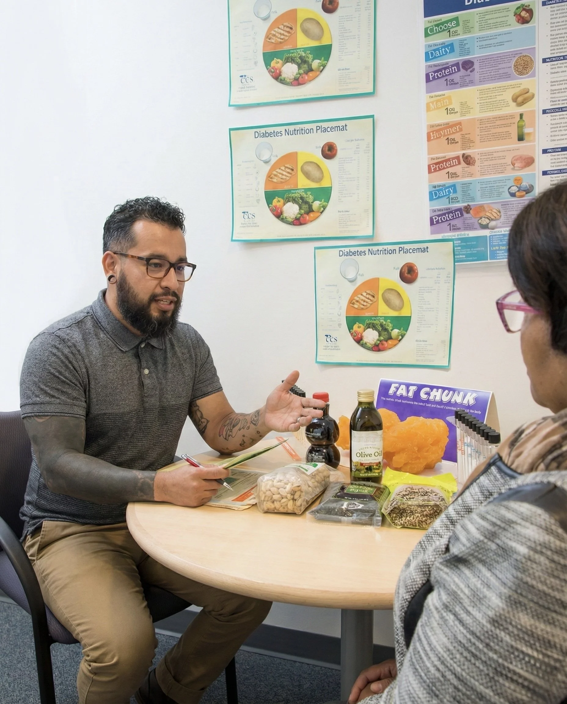 A man with glasses, a beard, and tattoos is talking to a woman with pink glasses at a round table. On the table, there are various food items including a bottle of olive oil, a package of nuts, a container of honey, and other snacks. The background features educational posters about nutrition and diabetes.