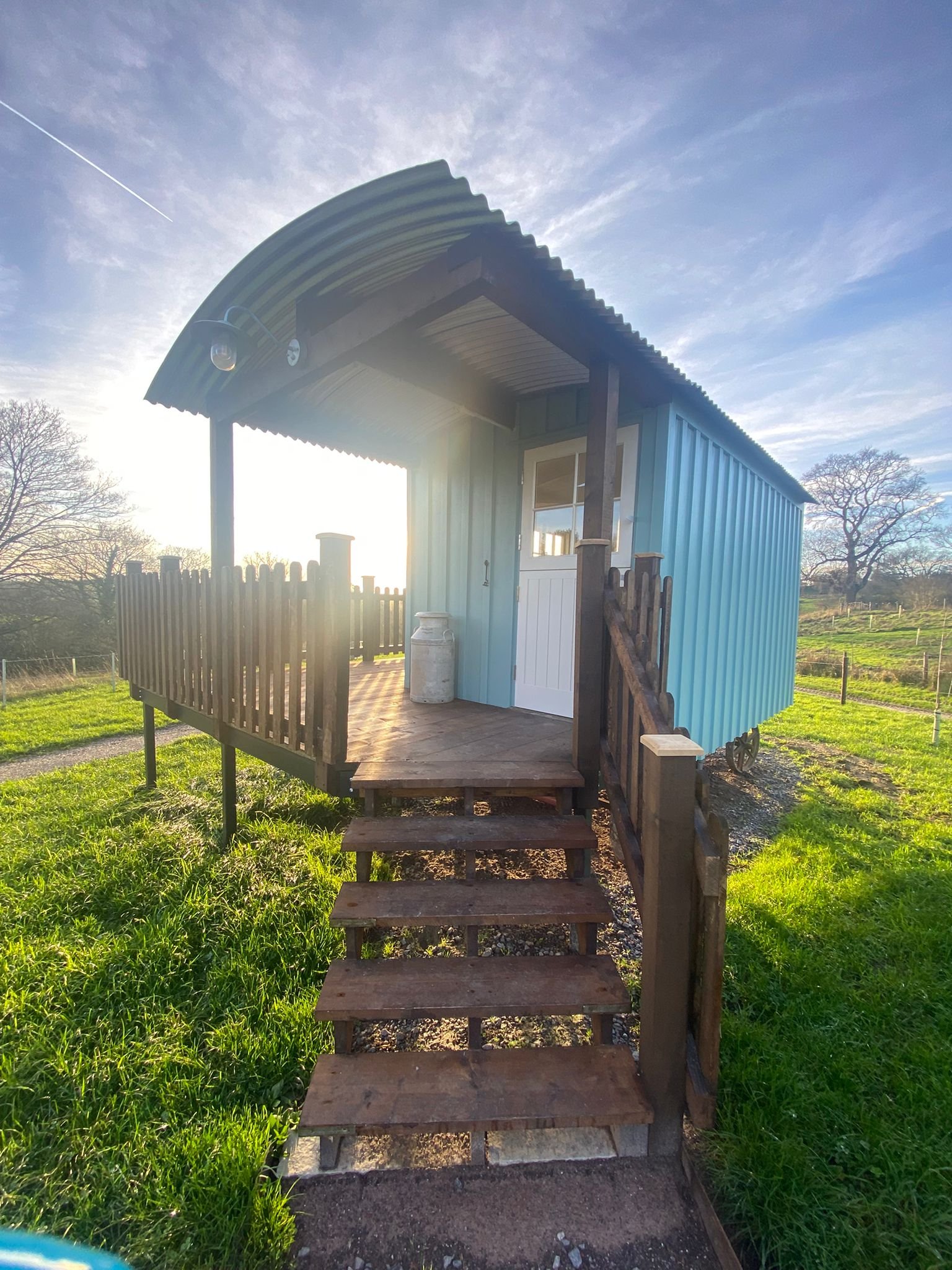 Small blue wooden shepherds hut with a corrugated metal roof, white door, and small porch with wooden railing, situated in a grassy outdoor area with trees and a clear sky in the background.
