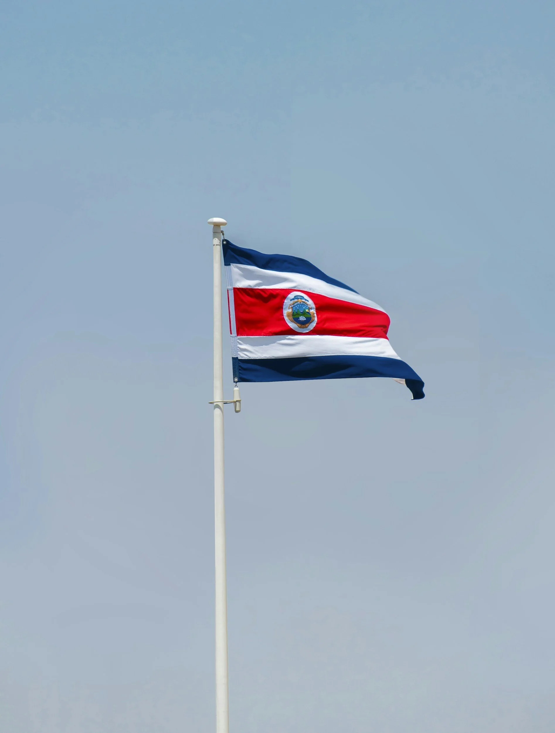 Costa Rican flag waving on a flagpole against a light blue sky.