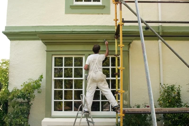 A person in white overalls painting the green trim of a house near a window with scaffolding nearby.
