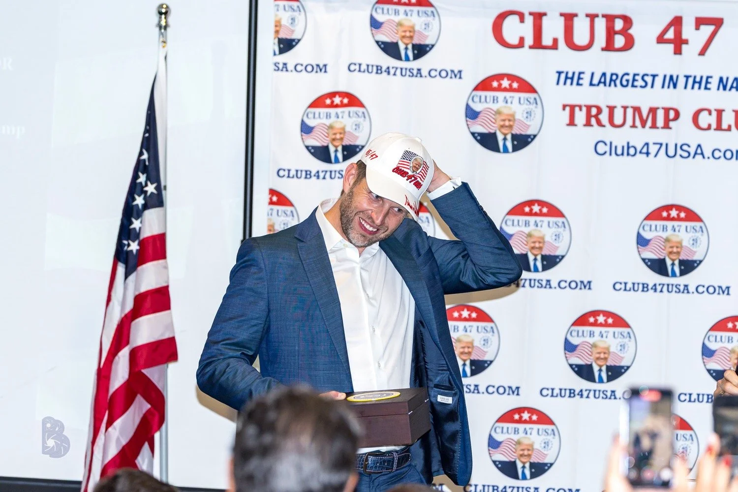 A man in a blue suit, white shirt, smiling, holding a box, touching his hat, standing in front of a backdrop with 'CLUB 47 USA' logos and American flags, at a patriotic event.