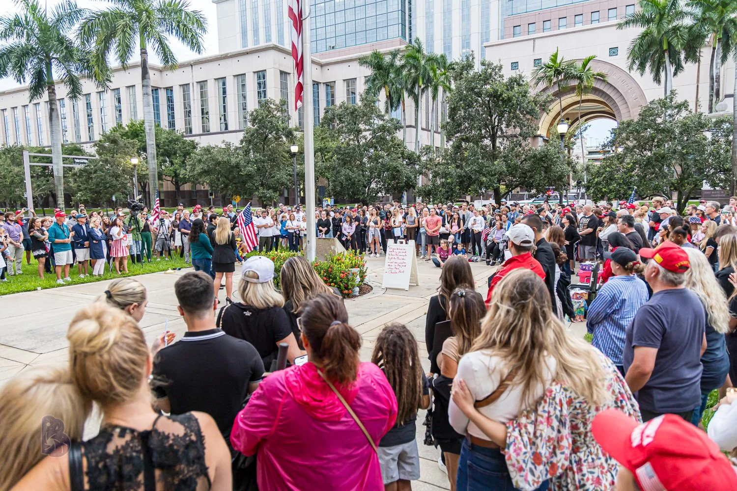 Crowd gathered at a political or protest rally in an urban park with green trees and high-rise buildings in the background, with one person speaking to the crowd near a flagpole and American flags.