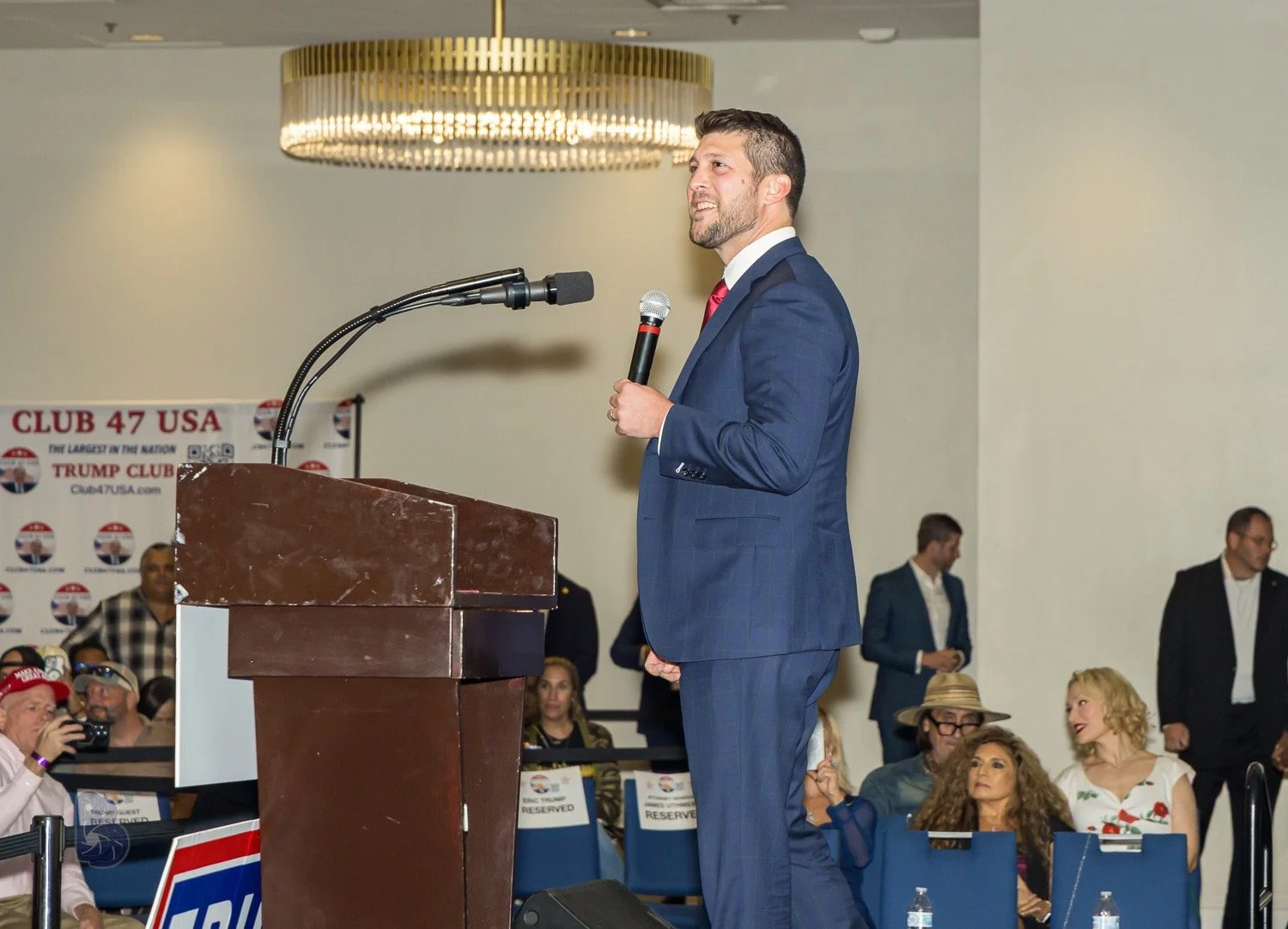 Man in a blue suit speaking into a microphone at a podium during a political event with a background banner that reads 'Club 47 USA' and 'Trump Club.' Audience members are seated, some wearing hats and sunglasses.