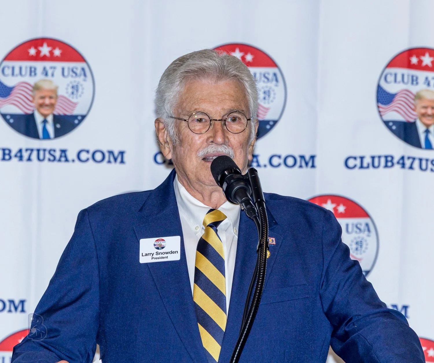 Older man with gray hair and glasses speaking into a microphone, wearing a blue suit, white shirt, and yellow striped tie, with a name badge that reads 'Larry Snowden President,' standing in front of a backdrop with 'Club 47 USA' and a logo featuring