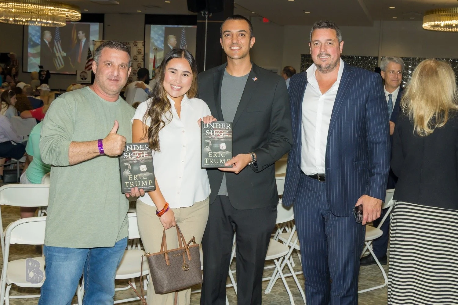 Group of five people posing at an event, with three holding copies of the book 'Undersiege' by Eric Trump, in a large room with screens and many seated attendees in the background.