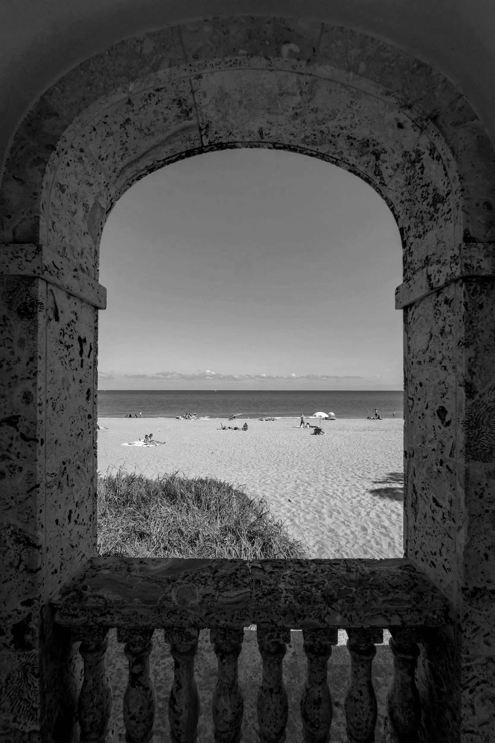 View of a beach framed by an arched stone window, with sand, people, umbrellas, and the ocean in the distance.