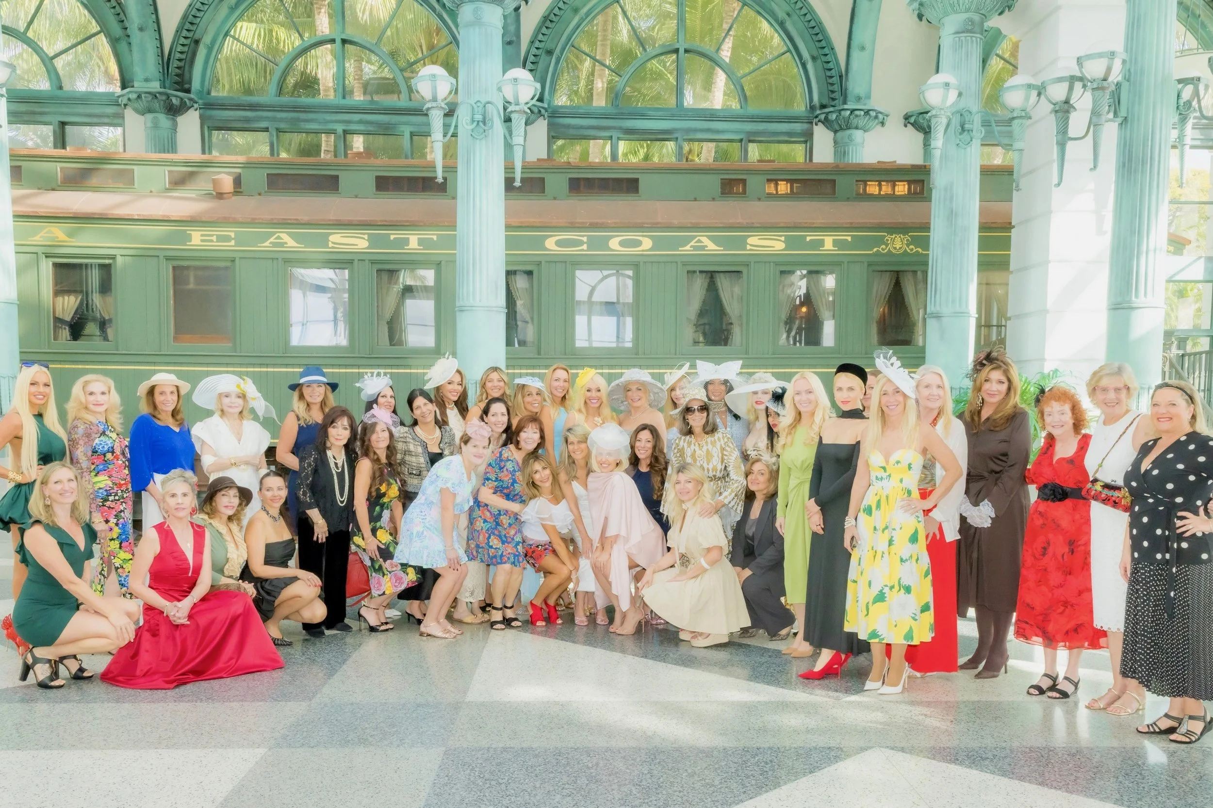 A large group of women dressed in colorful, elegant clothing and wide-brimmed hats pose together inside a historic train station with ornate green architecture and large windows.