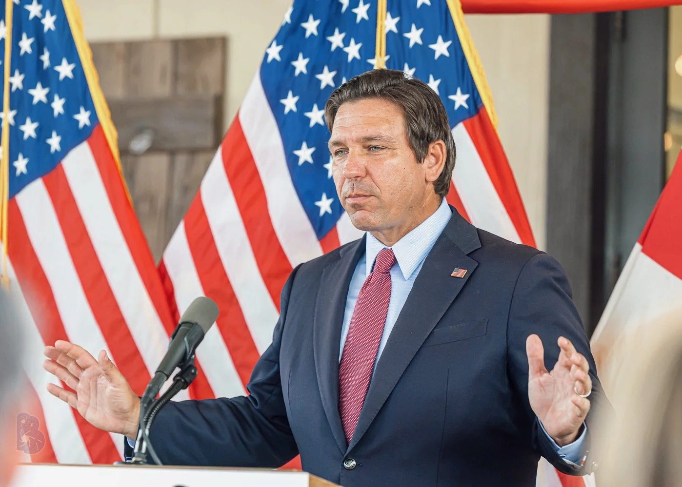A man in a suit and tie speaking at a podium, with American flags in the background.