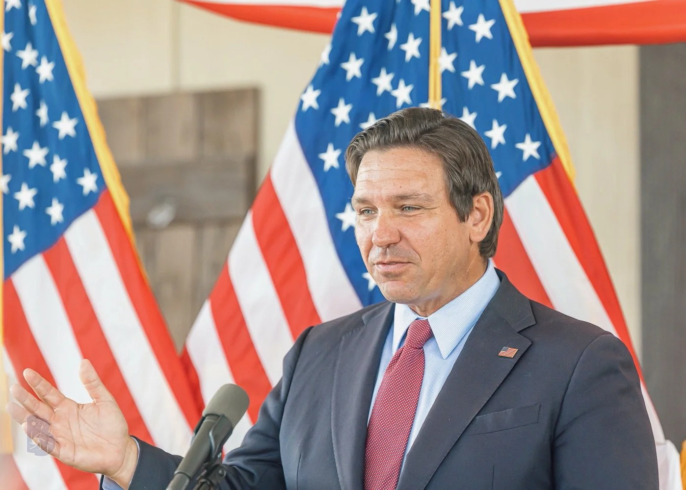 A man in a suit and red tie standing behind a microphone with American flags in the background, gesturing with his right hand.