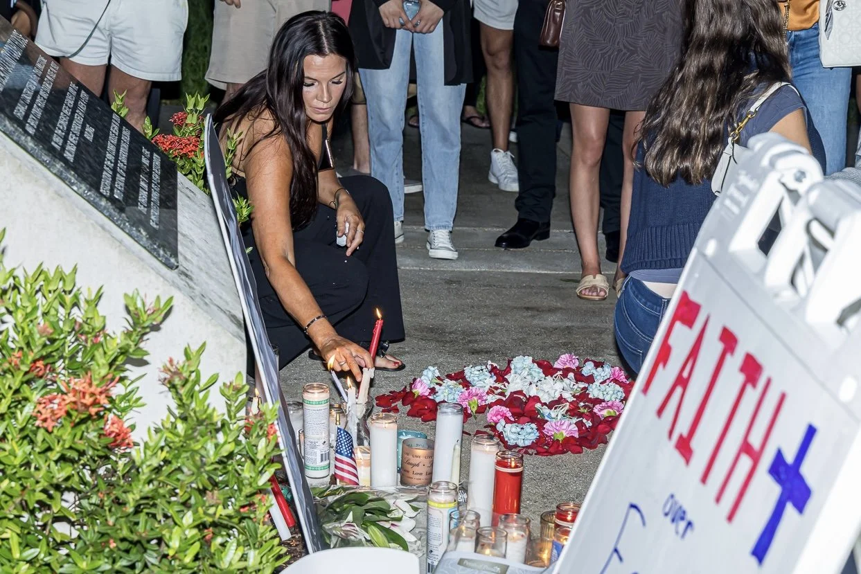 A woman is kneeling in front of a memorial with candles, flowers, and a sign that says 'FATHER' and 'OUR.' She appears to be placing something at the memorial during a gather of people.