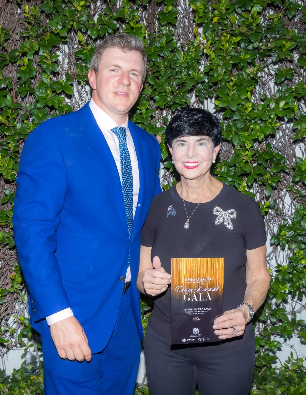 A man in a blue suit and a woman in a black shirt holding a program at an event titled 'Citizen Journalist Gala' with a background of green foliage.